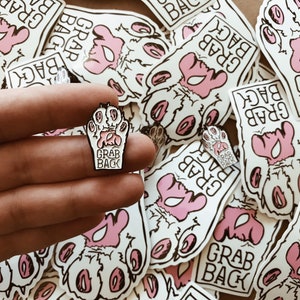 Pode incluir: Um broche de esmalte branco com uma pegada rosa e o texto "Grab Back" em letras pretas.