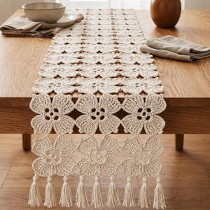 May include: A cream-coloured crochet table runner featuring a repeating floral motif pattern. The runner is constructed from interconnected flower shapes and is finished with a series of long, dangling tassels along the bottom edge.