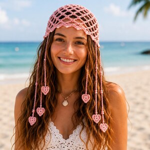Può includere: Un copricapo rosa all'uncinetto con un design a rete e fili pendenti adornati con decorazioni a forma di cuore. La persona indossa un top bianco e una collana di conchiglie.