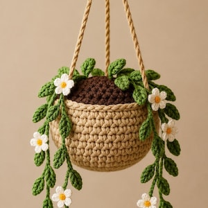 May include: A crocheted hanging basket with a light brown woven design. The basket holds a dark brown soil-like material and is adorned with green leaves and white flowers with yellow centers. The basket hangs from a tan rope.