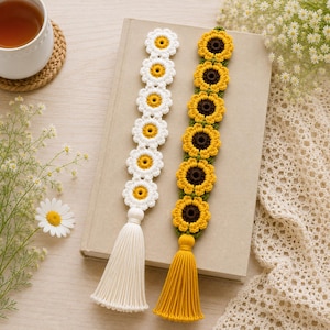 May include: Two handmade crochet bookmarks. One features white daisies with yellow centres and a white tassel. The other has yellow sunflowers with brown centres and a yellow tassel. Both are resting on a book.