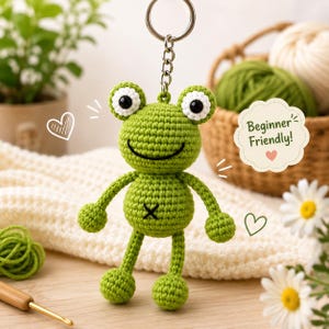 Pode incluir: Um chaveiro de sapo verde de crochê com um rosto sorridente, olhos pretos e um chaveiro prateado. O sapo tem um pequeno "X" preto na barriga. A imagem inclui o texto "Beginner Friendly!" e é cercada por fios e margaridas.