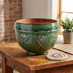 Peut inclure: Un grand pot de fleurs rond en céramique verte avec un motif sculpté détaillé. Le pot a une large ouverture et repose sur une table en bois. Une petite plante en pot et un dessous de verre sont également visibles.