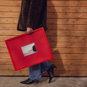 May include: A red picture frame containing a black and white photograph. The frame is held by a person wearing a brown leopard print coat, blue jeans, and black pointed-toe boots.