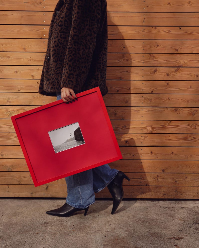May include: A red picture frame containing a black and white photograph. The frame is held by a person wearing a brown leopard print coat, blue jeans, and black pointed-toe boots.