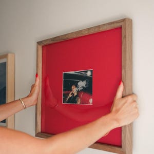 May include: A framed photograph with a wooden frame and a red background. The photograph is a black and white image of a person smoking in a car. The frame is being held by two hands with red nail varnish.