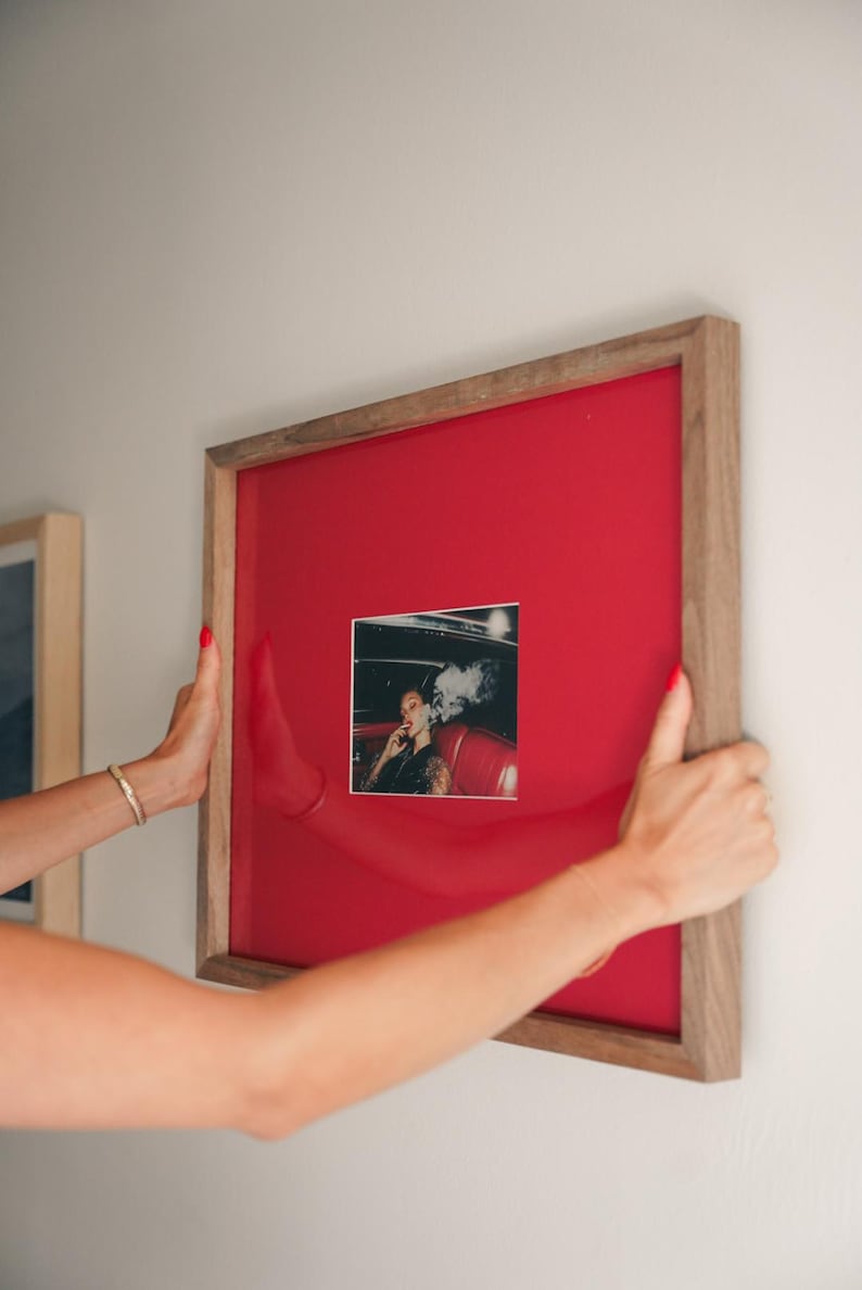 May include: A framed photograph with a wooden frame and a red background. The photograph is a black and white image of a person smoking in a car. The frame is being held by two hands with red nail varnish.
