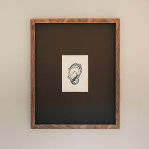 May include: A framed black and white oyster shell drawing. The artwork is centred on a dark brown background within a rectangular wooden frame. The frame has a natural wood grain finish.