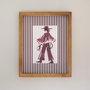 May include: Framed art print. The burgundy illustration depicts a person in a hat and cape, holding a rope. The artwork is set against a striped background of white and burgundy lines, within a wooden frame.