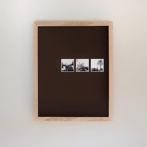 May include: A wooden-framed display with three small black and white photographs. The background is dark brown. The photographs show outdoor scenes. The frame is made of light-coloured wood.