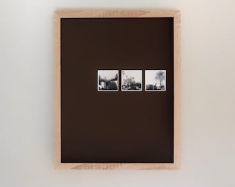 May include: A framed display featuring three small black and white photographs. The frame is made of light-colored wood, and the background is a dark brown. The photographs depict outdoor scenes.
