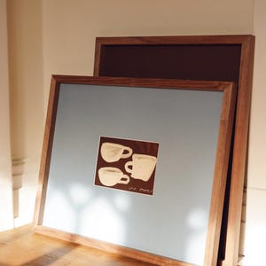 K&ouml;nnte beinhalten: Zwei gerahmte Kunstdrucke, die an einer Wand lehnen. Der vordere Druck hat einen hellblauen Hintergrund mit einem kleinen quadratischen Bild von drei wei&szlig;en Kaffeetassen. Die Rahmen sind aus Holz.