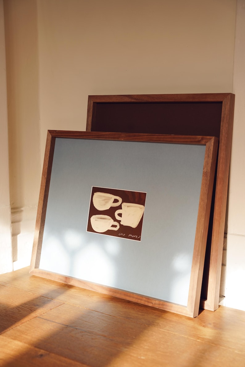 K&ouml;nnte beinhalten: Zwei gerahmte Kunstdrucke, die an einer Wand lehnen. Der vordere Druck hat einen hellblauen Hintergrund mit einem kleinen quadratischen Bild von drei wei&szlig;en Kaffeetassen. Die Rahmen sind aus Holz.