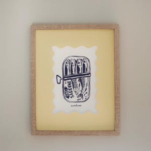 May include: Framed print with a navy blue illustration of sardines in a tin. The artwork is set against a yellow background with a white, scalloped border. The frame is light wood-coloured.