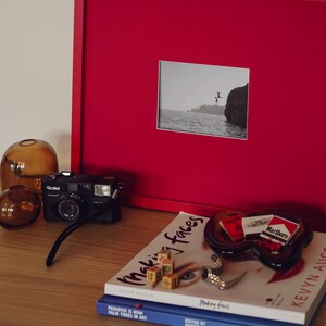 May include: A collection of objects on a wooden surface, including a red framed picture with a monochrome image, a black camera, and glass ornaments. Books and an ashtray with cigarette packs complete the arrangement.