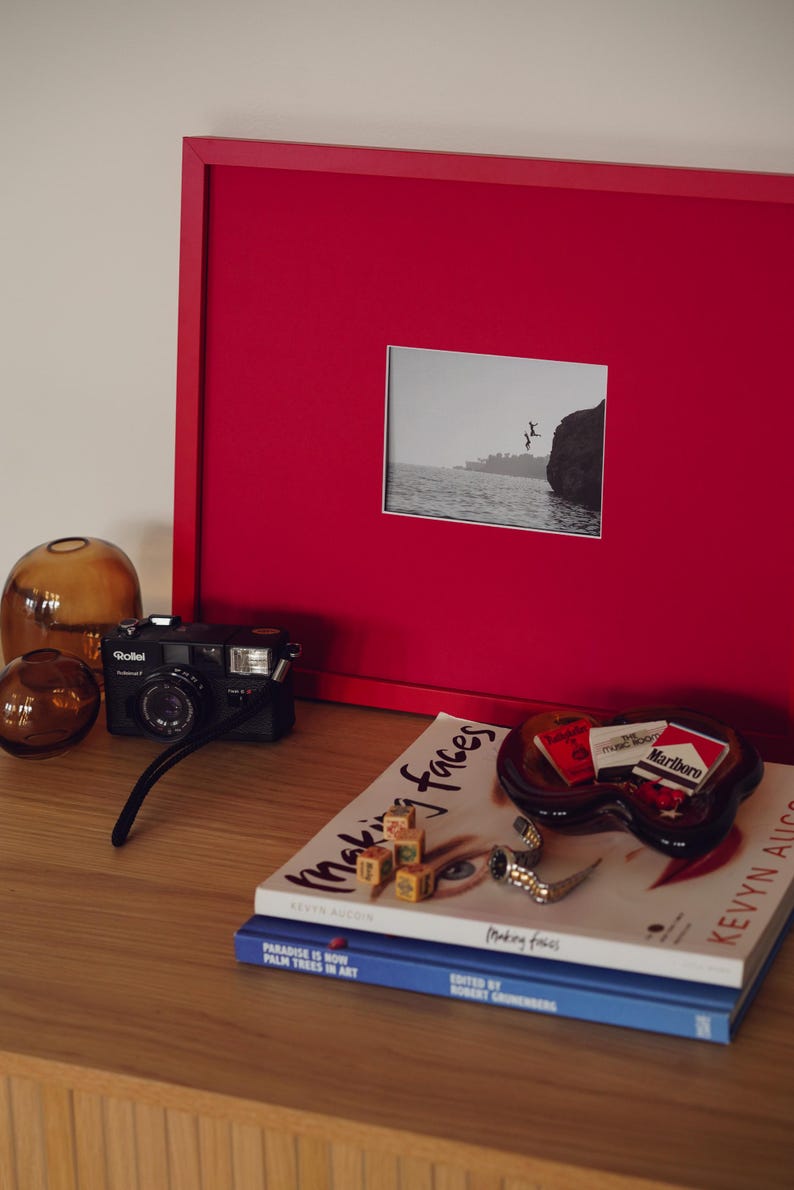 May include: A collection of objects on a wooden surface, including a red framed picture with a monochrome image, a black camera, and glass ornaments. Books and an ashtray with cigarette packs complete the arrangement.