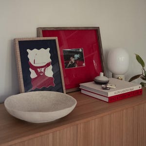 May include: A wooden cabinet displaying a light-coloured, round bowl, framed artwork, books, and a white lamp. The artwork includes a red and white image and a photograph. The books are titled "Slim Aarons Style" and "Vogue Paris 100 Years".