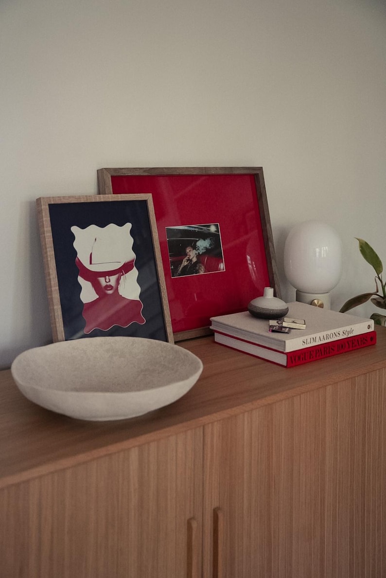 May include: A wooden cabinet displaying a light-coloured, round bowl, framed artwork, books, and a white lamp. The artwork includes a red and white image and a photograph. The books are titled "Slim Aarons Style" and "Vogue Paris 100 Years".