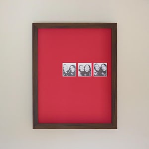 Peut inclure: Une œuvre d'art encadrée avec un fond rouge vif. Trois petites photos de style photomaton en noir et blanc sont fixées sur la surface rouge. Le cadre est de couleur marron foncé.