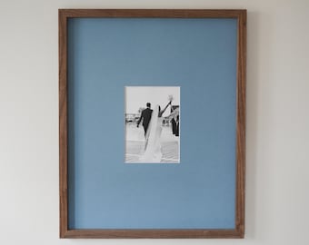 May include: A framed photograph featuring a black and white image of a wedding couple. The photograph is set against a light blue background within a wooden frame. The couple is walking away from the camera, with the bride raising her bouquet.