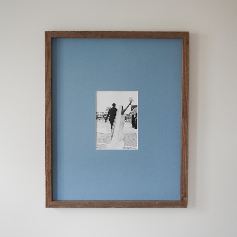 K&ouml;nnte beinhalten: Gerahmtes Foto mit einem Schwarz-Wei&szlig;-Bild eines Hochzeitspaares. Das Foto befindet sich vor einem hellblauen Hintergrund in einem Holzrahmen. Das Paar entfernt sich von der Kamera, die Braut hebt ihren Brautstrau&szlig;.