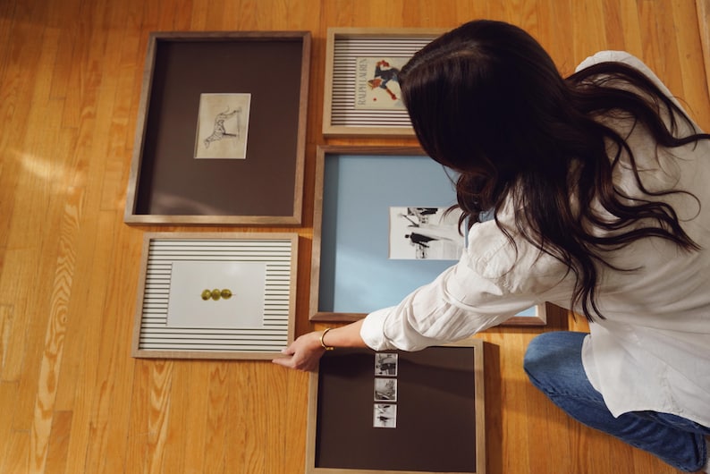 K&ouml;nnte beinhalten: Eine Sammlung gerahmter Kunstwerke in verschiedenen Gr&ouml;&szlig;en und Stilen. Die Rahmen sind in Braun- und Naturholzt&ouml;nen gehalten. Die Kunstwerke umfassen Zeichnungen und Illustrationen. Die St&uuml;cke sind auf einer Holzoberfl&auml;che angeordnet.