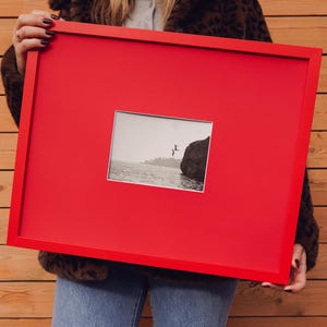 May include: A bright red picture frame holding a black and white photograph of a person leaping into the sea. The frame is rectangular and is being held. The photograph is centrally positioned within the frame.