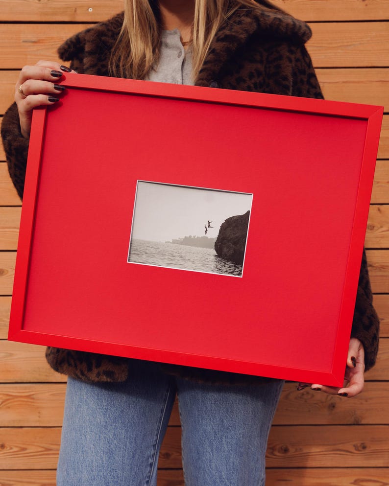 May include: A bright red picture frame holding a black and white photograph of a person leaping into the sea. The frame is rectangular and is being held. The photograph is centrally positioned within the frame.