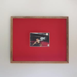 May include: Framed print with a photograph of a person in a car, exhaling smoke. The print is set against a bright red background within a wooden frame. The image captures a moment of leisure.