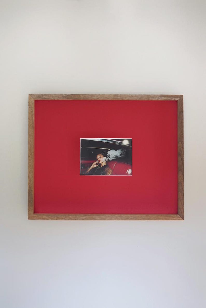 May include: Framed print with a photograph of a person in a car, exhaling smoke. The print is set against a bright red background within a wooden frame. The image captures a moment of leisure.