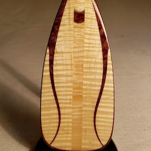 May include: A wooden paddle with a natural wood finish and dark brown accents. The paddle has a unique grain pattern and is displayed on a black stand.