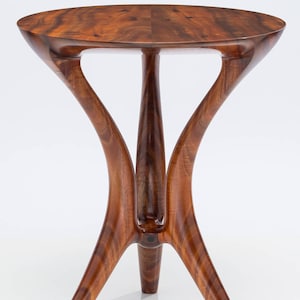 May include: A round wooden table with a unique, sculptural base. The table has a dark brown wood finish and a smooth, polished surface. The base is made of three curved legs that meet in the center. The table is a beautiful example of handcrafted furniture.