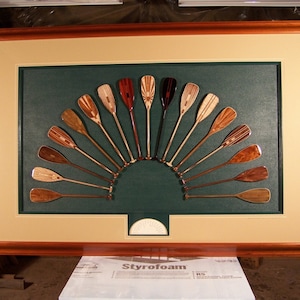 May include: A framed artwork featuring a semi-circular arrangement of wooden paddles. The paddles, in various wood tones, are set against a dark green background, framed with a light beige mat and a dark brown wooden frame. The artwork is displayed on a white surface.