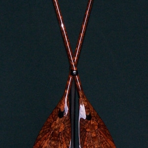 May include: Two wooden paddles with intricate wood grain patterns and black accents are crossed and displayed on a black stand.