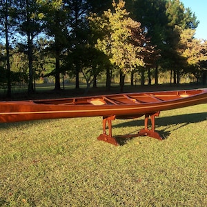 May include: A wooden canoe with a natural wood finish and a dark brown stain. The canoe is resting on a wooden stand and is surrounded by green grass.