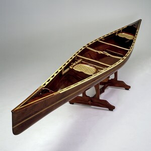 May include: A wooden canoe with a dark brown stain and light wood trim. The canoe is on a wooden stand with four legs.