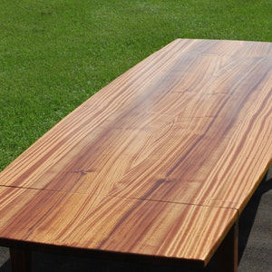 May include: A long, wooden table with a natural wood finish. The table top is made of multiple planks of wood that have been joined together. The table has a slightly curved edge and is supported by four legs.