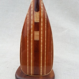 May include: A handcrafted wooden paddle with a dark brown base. The paddle features alternating vertical stripes of light and dark wood, creating a visually appealing design. The paddle's shape is elongated and symmetrical, with a rounded tip.