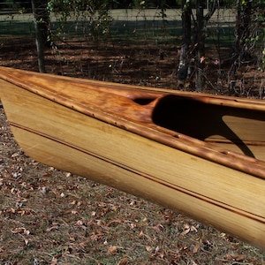 May include: A wooden canoe with a natural wood finish. The canoe has a light brown color with dark brown accents. The canoe is sitting on a bed of brown leaves.