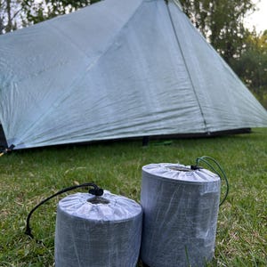 May include: Two cylindrical, gray stuff sacks with drawstring closures sit on green grass in front of a light green tent. The stuff sacks have black and green drawstrings. The tent is pitched in a grassy area.