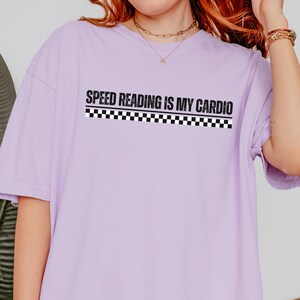 Peut inclure: Un t-shirt à manches courtes violet clair avec le texte noir "SPEED READING IS MY CARDIO" imprimé au-dessus d'un motif à damier noir et blanc. Le vêtement présente une encolure ronde et une coupe décontractée, avec le motif centré sur la poitrine.