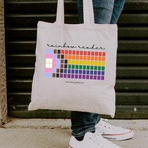 May include: A cream-colored canvas tote bag featuring the text "rainbow reader" in black script above a grid of colorful squares arranged in a rainbow pattern. Below the grid, the text "THE CASUAL BOOKCLUB" is printed in small black letters.