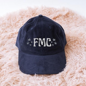 May include: Navy blue baseball cap with the letters "FMC" embroidered in white, flanked by stars. The cap is made of a soft material and has a curved brim. The hat is displayed on a peach-colored, fluffy surface.