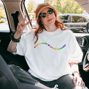 May include: White t-shirt with the rainbow text "STRAIGHT IS OVERRATED". The shirt is worn by a person in a car, wearing sunglasses and a brown hat. The person is giving a peace sign.