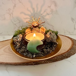 May include: A gold-toned metal tray holding a lit tea light candle, a green crescent moon-shaped stone, metallic pyrite clusters, light-coloured crystals, and green moss. A gold sun-shaped ornament is positioned behind the candle holder.