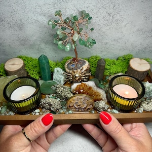 May include: A rectangular wooden tray containing a wire-wrapped green crystal tree, two lit tea light candles in ribbed green glass holders, various raw crystals including citrine and pyrite, green moss, two small wood slices, and a circular token.