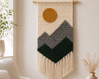 May include: A rectangular crochet wall hanging featuring a landscape design with a mustard yellow sun, grey mid-ground mountains, and dark green foreground mountains. The piece is suspended from a wooden dowel with a cream cord and has fringe.