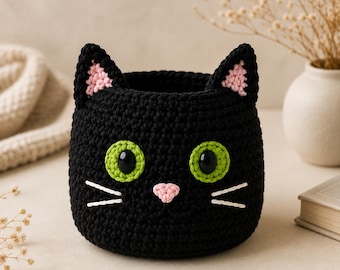 May include: A black crocheted basket shaped like a cat head. It features two pointed ears with pink inner details, large bright green eyes with black pupils, a small pink nose, and three white stitched whiskers on each side of the face.