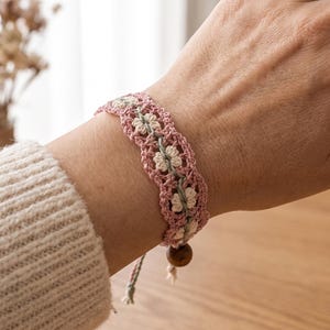 Pode incluir: Uma pulseira de crochê com um padrão floral repetitivo em fio rosa suave e creme, acentuado com um detalhe central em verde sálvia. A pulseira é finalizada com uma conta de madeira castanha na extremidade do fecho ajustável.