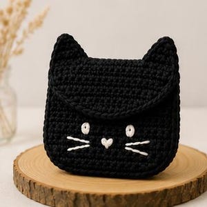 May include: A black crocheted pouch shaped like a cat head with pointed ears. The front features white embroidered details including two circular eyes, a small heart-shaped nose, and three whiskers on each side of the face, creating a feline face.